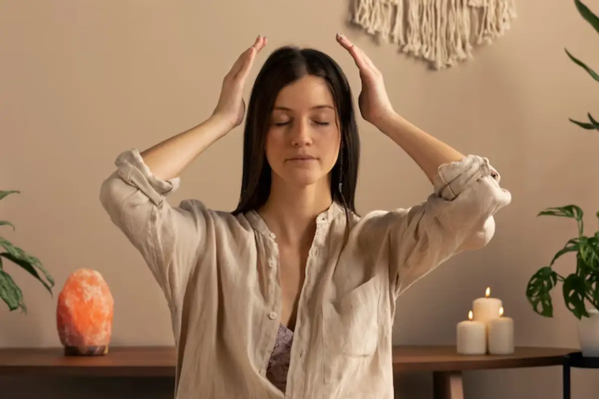 A woman in a beige shirt is seated with her hands raised, surrounded by plants, candles, and a decorative lamp in a serene setting.