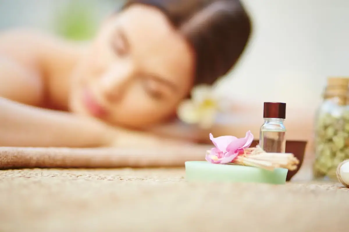 A relaxing spa setting featuring a green soap, essential oil bottle, and a pink flower on a textured surface, evoking tranquility.