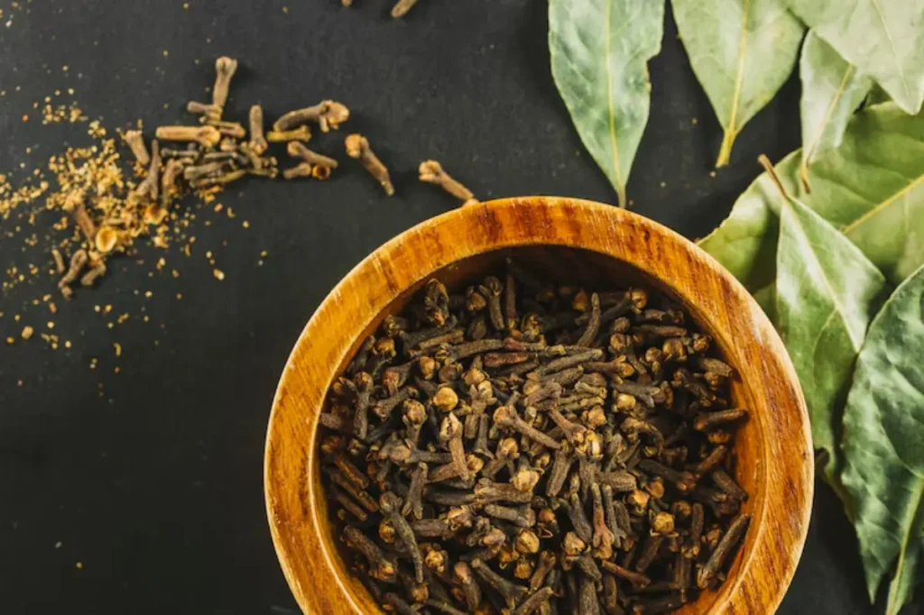 A wooden bowl filled with whole cloves, surrounded by scattered cloves and bay leaves, set against a dark background.