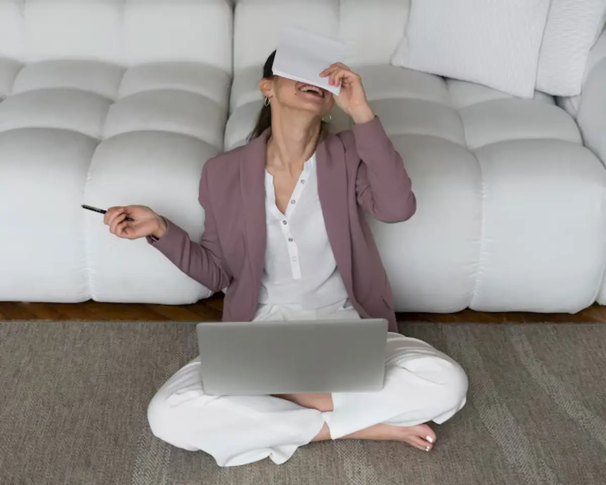 A person sitting cross-legged on a rug with a laptop, holding a pen, appearing thoughtful in a modern living room setting.