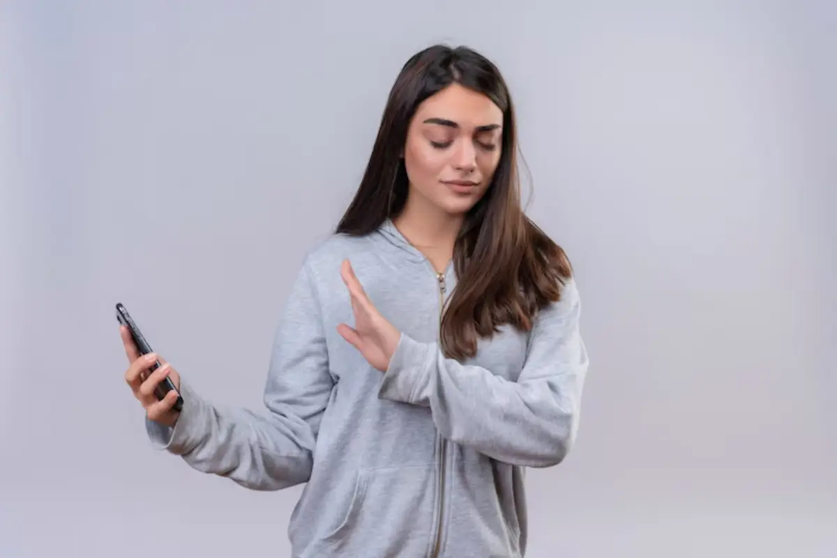 A young woman in a gray hoodie holds a smartphone in one hand and gestures with the other, against a light gray background.