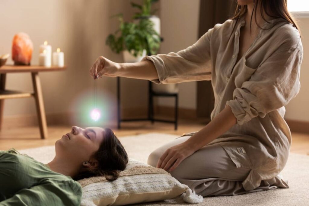 A practitioner conducts a healing session, holding a pendulum above a person lying on a cushion, surrounded by calming decor.