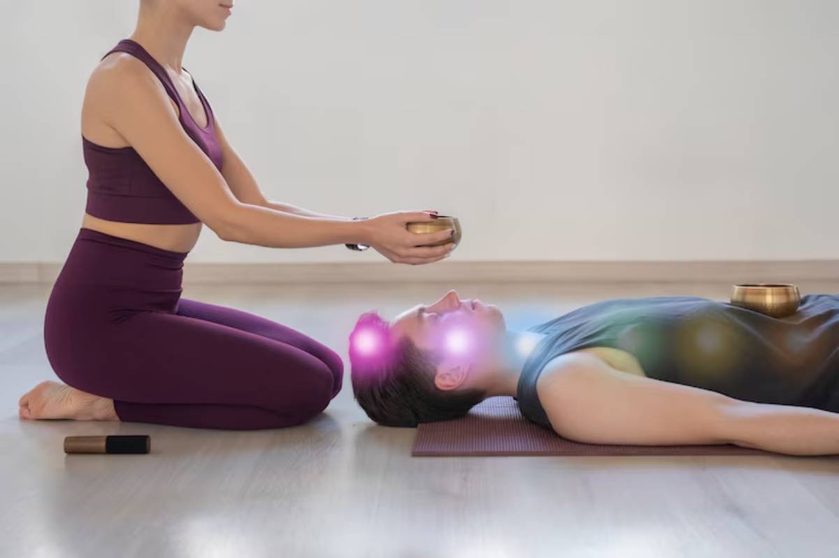 A woman in a purple outfit performs a healing practice, holding a bowl above a person lying on a yoga mat in a serene space.