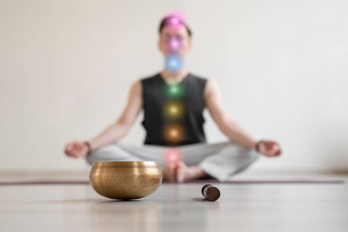 A person sits cross-legged on a yoga mat, meditating with a singing bowl and mallet in front, surrounded by colorful chakra lights.