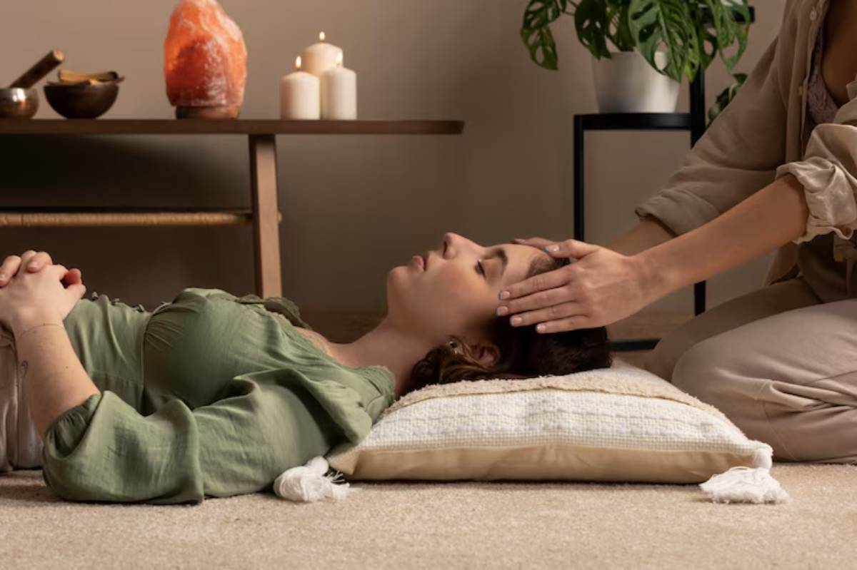 A person lies on a pillow while another gently places their hand on her head in a peaceful, ambient setting with candles and plants.