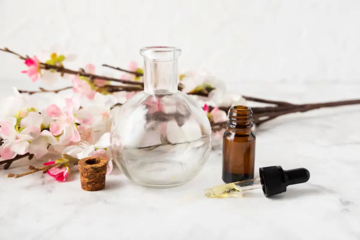 A clear glass bottle and a dark amber dropper bottle on a marble surface, surrounded by delicate pink cherry blossom branches.