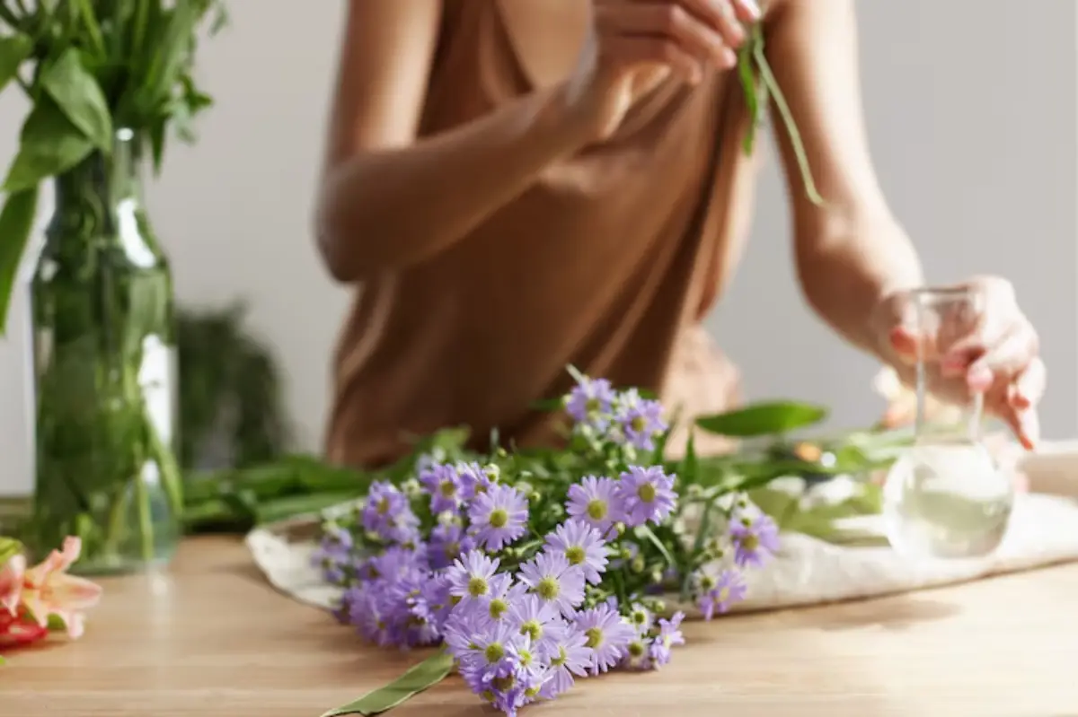 A person arranging purple flowers on a wooden table, holding a stem while preparing a vase with water nearby.