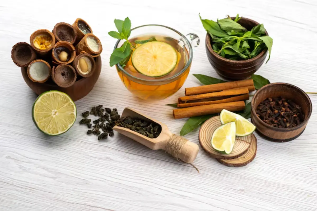 A collection of spices and herbs including green tea, cinnamon sticks, and lime, alongside a cup of yellow tea with a lemon slice.