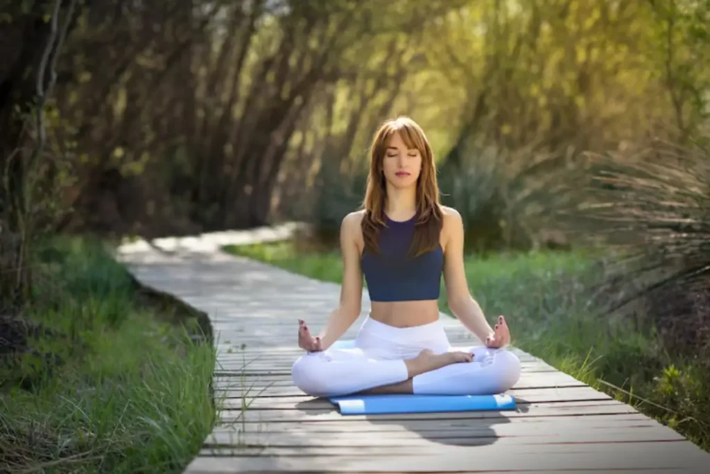 A woman meditates peacefully on a blue mat surrounded by greenery on a wooden path in a serene outdoor setting.