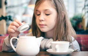 A person in a cozy café pours from a white teapot into a cup, surrounded by warm lighting and hints of greenery.