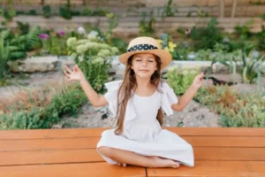 A girl in a white dress and a straw hat sits cross-legged on a wooden deck, surrounded by vibrant garden plants, forming a peace sign.