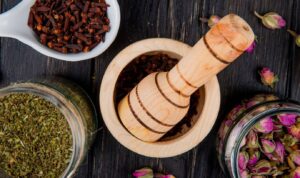A wooden mortar and pestle surrounded by jars of dried herbs, cloves, and rosebuds on a dark wooden surface.