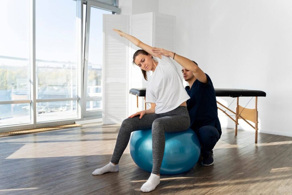A person performs an exercise on a blue stability ball, assisted by a trainer in a bright, spacious room with large windows.