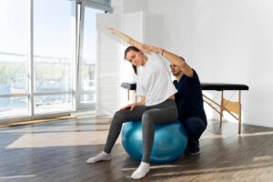 A person performs an exercise on a blue stability ball, assisted by a trainer in a bright, spacious room with large windows.