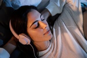 A person lies comfortably on a couch, wearing white headphones, surrounded by cozy blankets and soft pillows, lost in music.