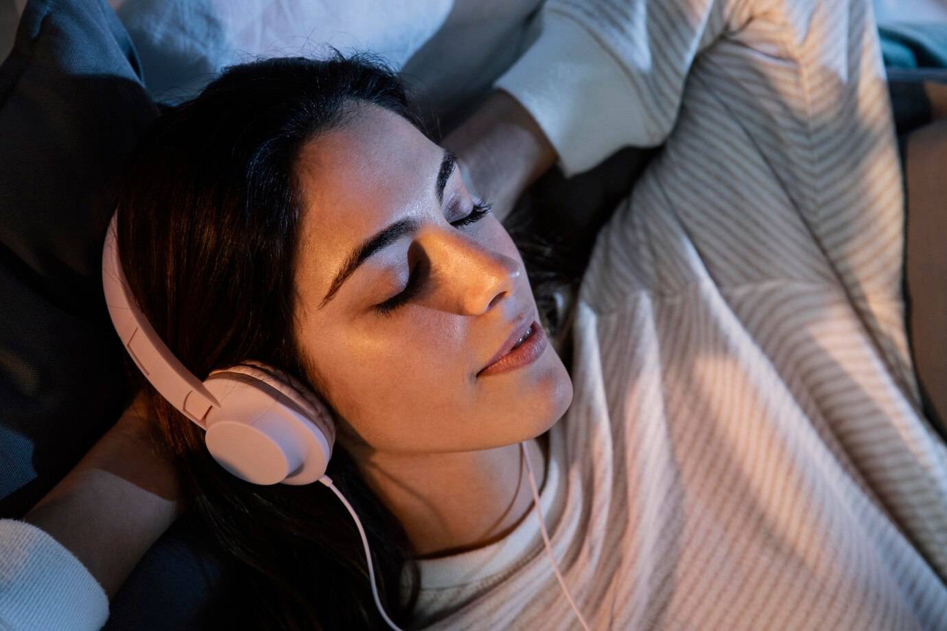 A person lies comfortably on a couch, wearing white headphones, surrounded by cozy blankets and soft pillows, lost in music.