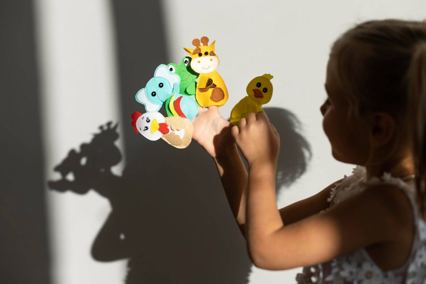 A hand displays colorful felt finger puppets of various animals, casting playful shadows on a textured wall in soft sunlight.