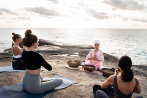 A serene outdoor yoga session at sunset, featuring participants meditating with singing bowls on a rocky coastline.
