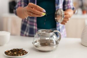 A person adds loose tea leaves into a glass teapot with a metal infuser, beside a plate of dried tea leaves.