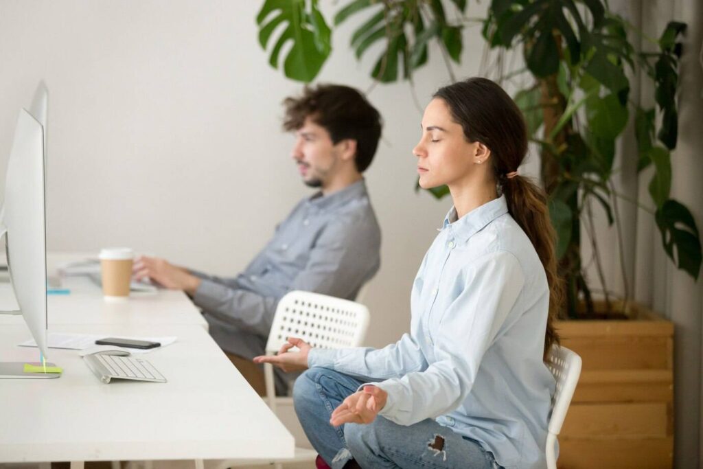 A woman meditates at a desk, while a man works on a computer, with indoor plants adding a calming atmosphere.