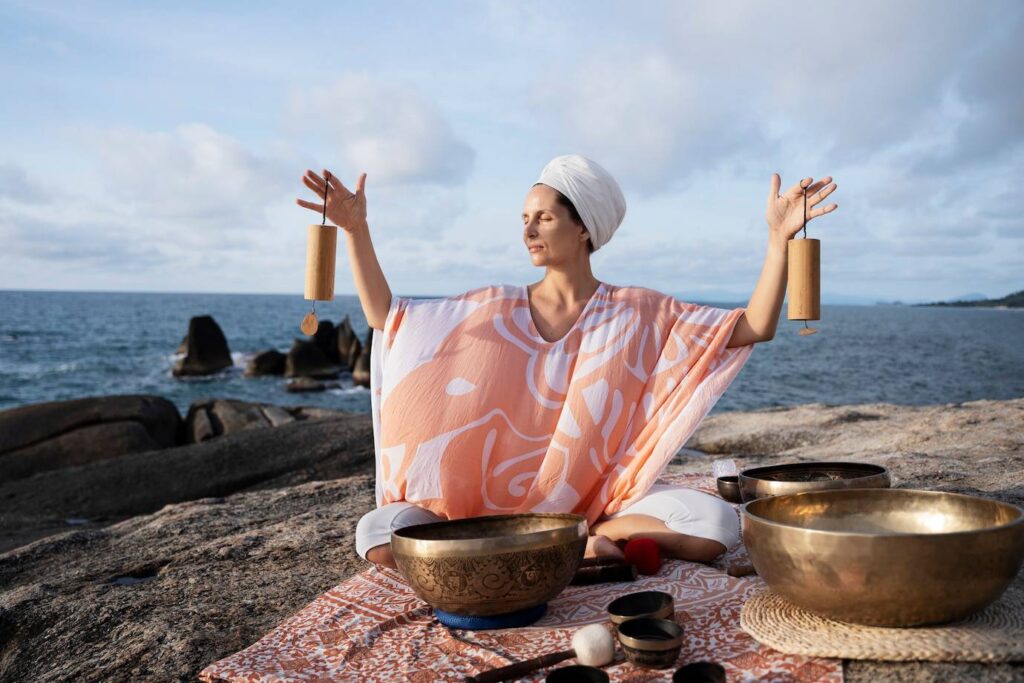 A person in a vibrant orange and white poncho holds sound bowls, creating a serene ambiance by the ocean.