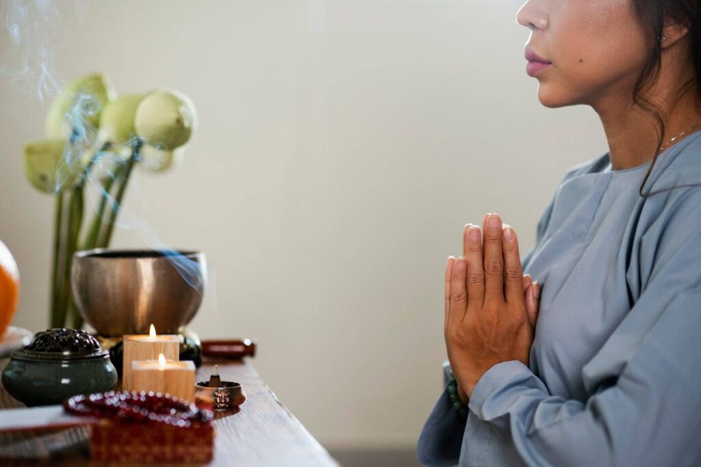 A serene setting with a person in prayer, surrounded by candles, incense smoke, and a bowl, alongside a vase of fresh lotus flowers.