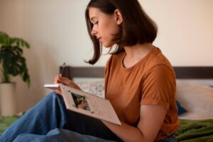 A young woman in an orange shirt sits on a bed, writing in a notebook with a plant visible in the background.