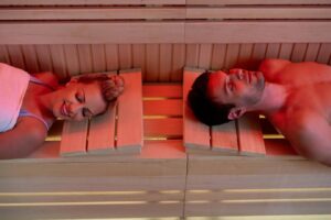 Two people resting on wooden benches inside a sauna, surrounded by warm, softly illuminated wooden walls.