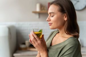 A person holds a glass of yellow beverage with mint leaves, standing in a bright kitchen with modern decor.