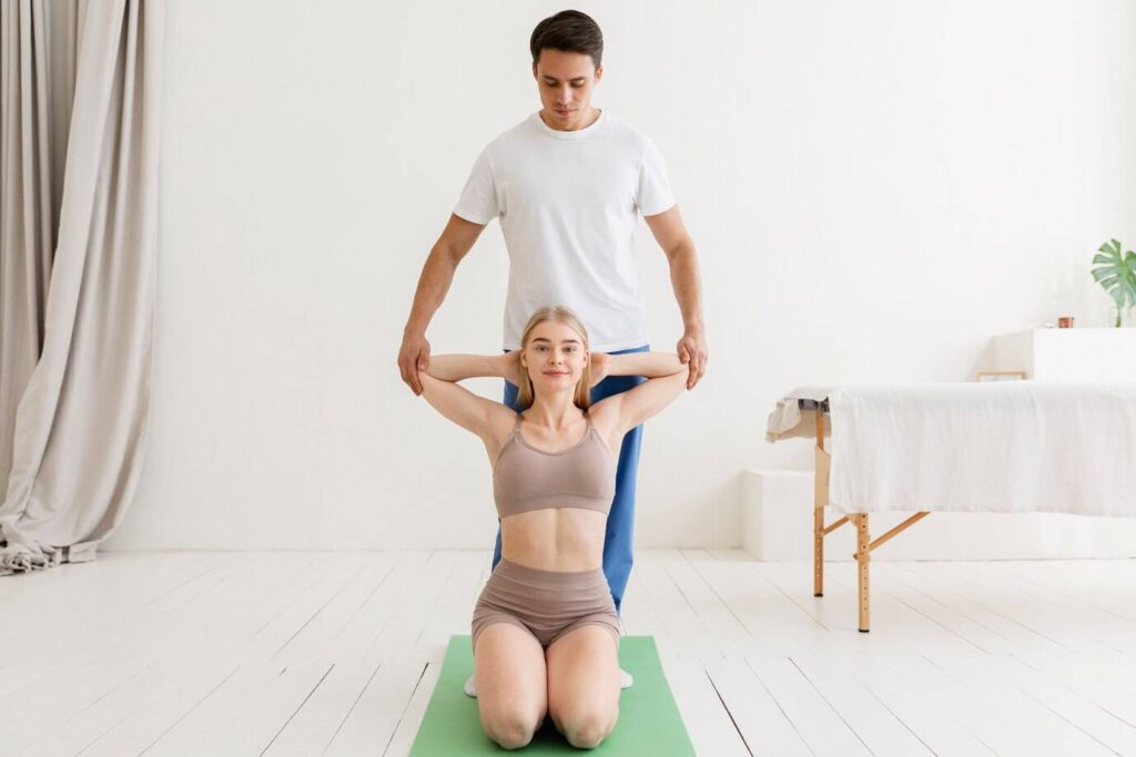A person assists another in a posture on a yoga mat in a bright, minimalistic studio. White walls and soft furnishings create a calm atmosphere.