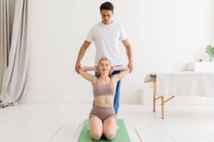 A person assists another in a posture on a yoga mat in a bright, minimalistic studio. White walls and soft furnishings create a calm atmosphere.