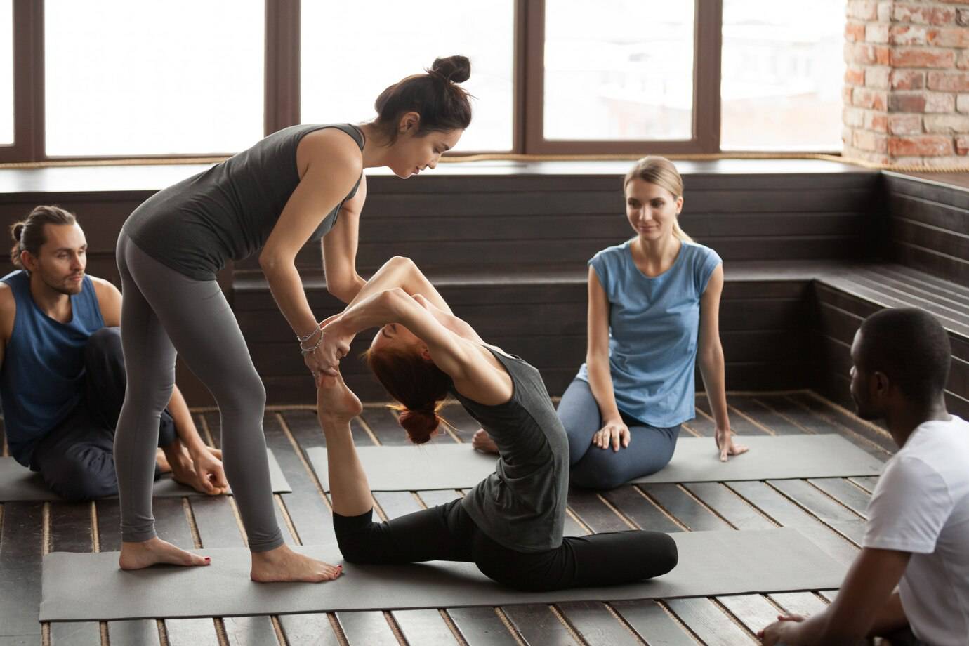 A group of individuals engages in a yoga class, practicing poses and providing assistance in a serene, well-lit studio space.