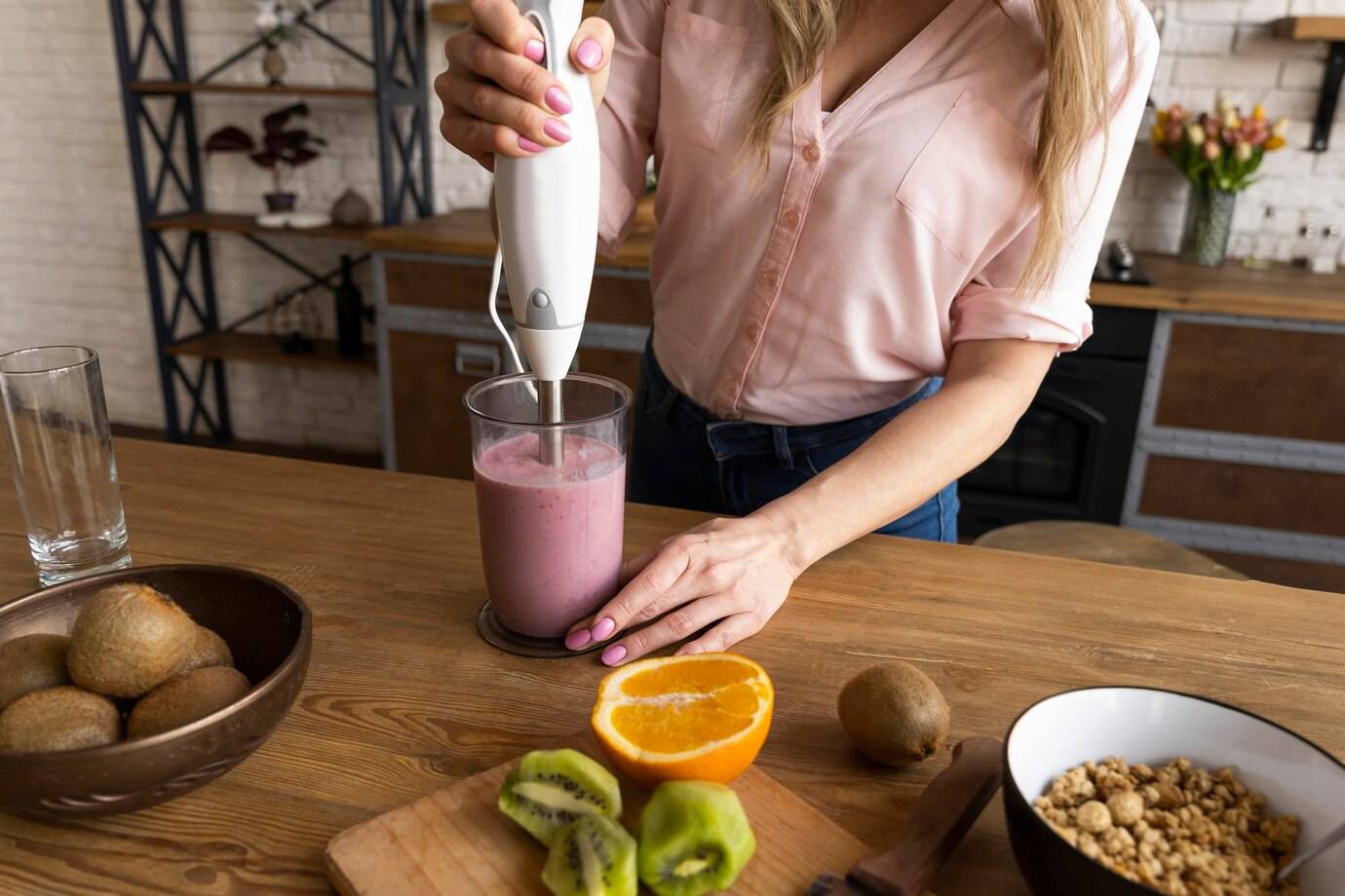 A woman in a pink shirt uses an immersion blender in a glass with pink smoothie, surrounded by fruits and a bowl of granola.