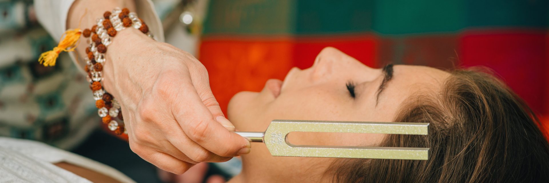 A hand holds a metal tool above a person's forehead, accompanied by a colorful background and a beaded bracelet on the wrist.