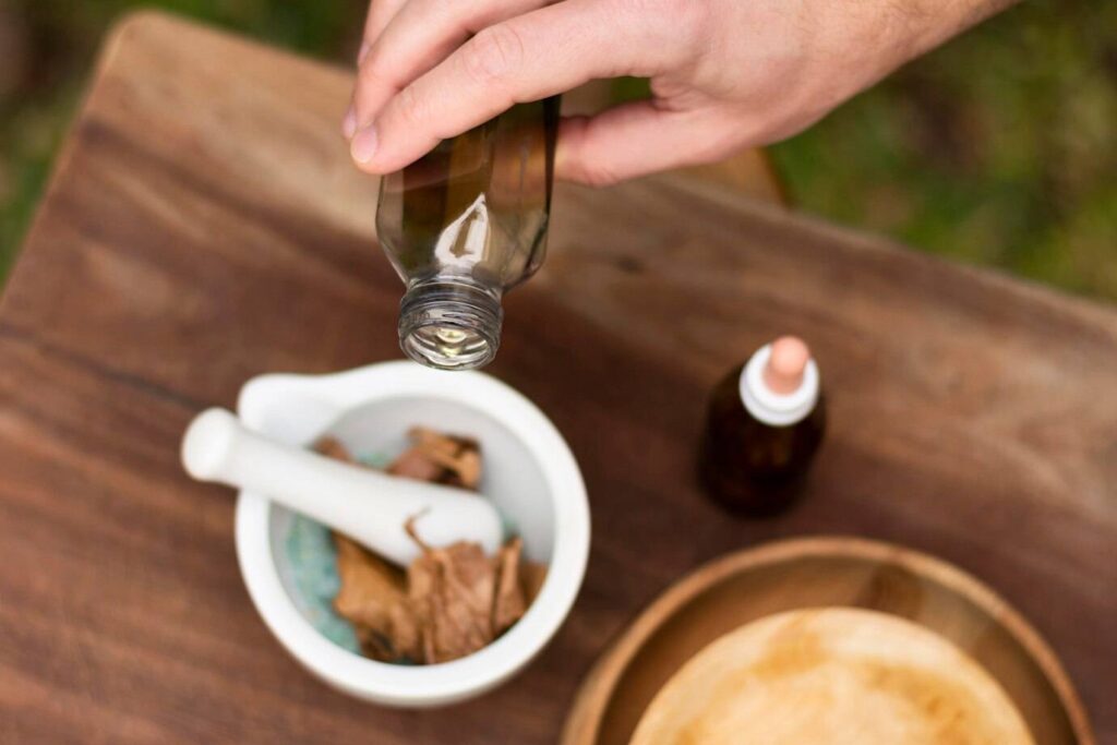 A hand pours oil from a glass bottle into a white mortar containing brown ingredients, with a small dropper bottle in the background.