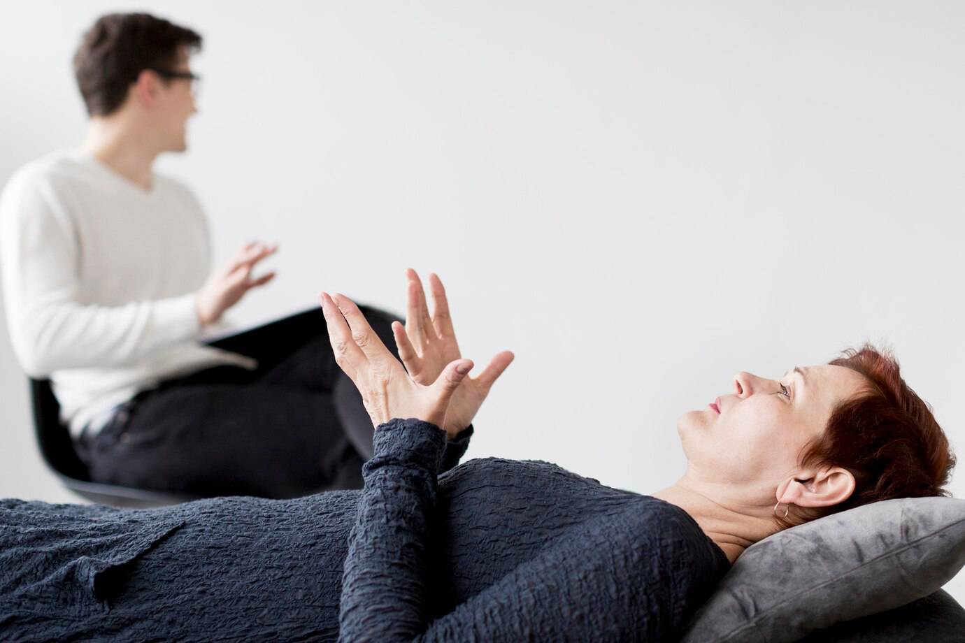 A woman lies on a couch expressing herself, while a man sits in a chair, engaged in conversation, in a serene, minimalistic setting.