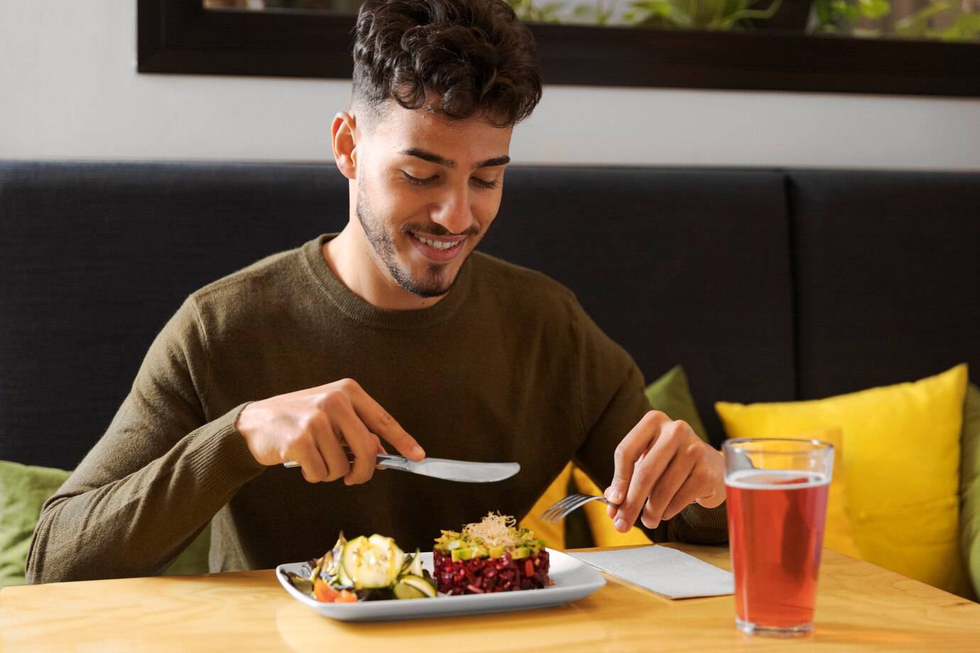 A person in a brown sweater holds a knife and fork, preparing to eat a colorful vegetable dish, with a drink beside them.