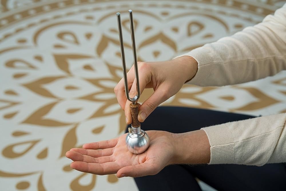 A hand holds a silver tuning fork above an open palm, set against a decorative mandala-patterned floor.