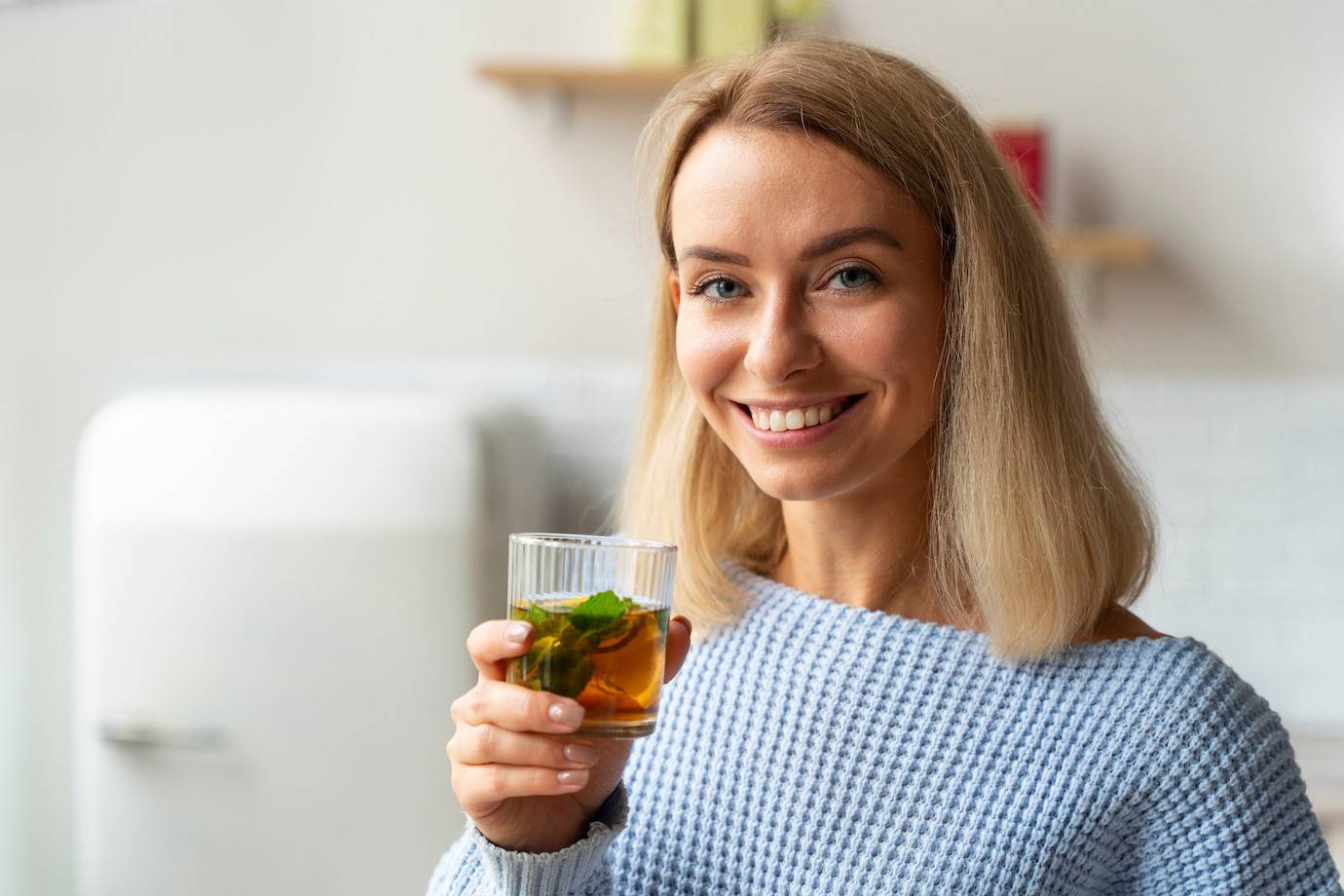 A person in a light blue sweater holds a glass of beverage with mint leaves, standing in a bright kitchen setting.