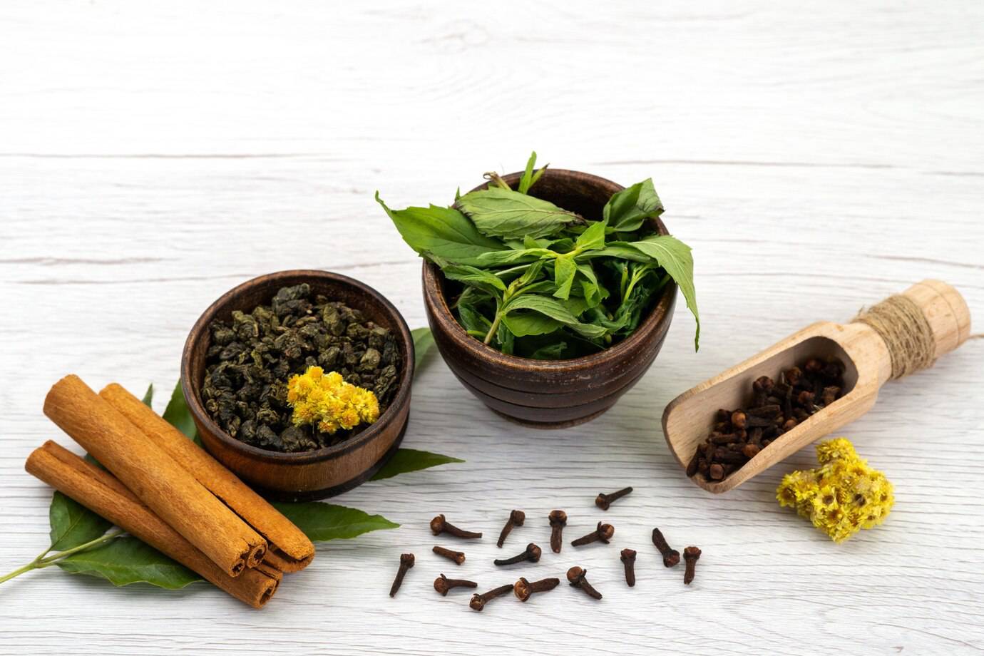 A wooden bowl with green tea leaves, another with dried leaves, cinnamon sticks, cloves, and yellow flowers on a light wood surface.