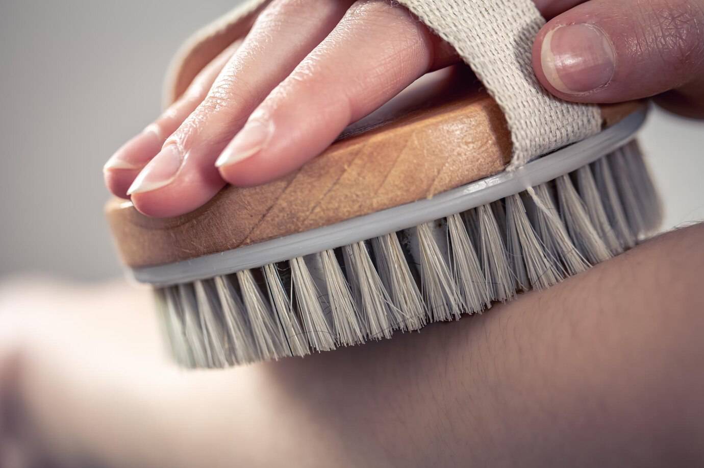 A close-up of a hand holding a wooden body brush, gently brushing a person's skin, emphasizing skincare and relaxation.