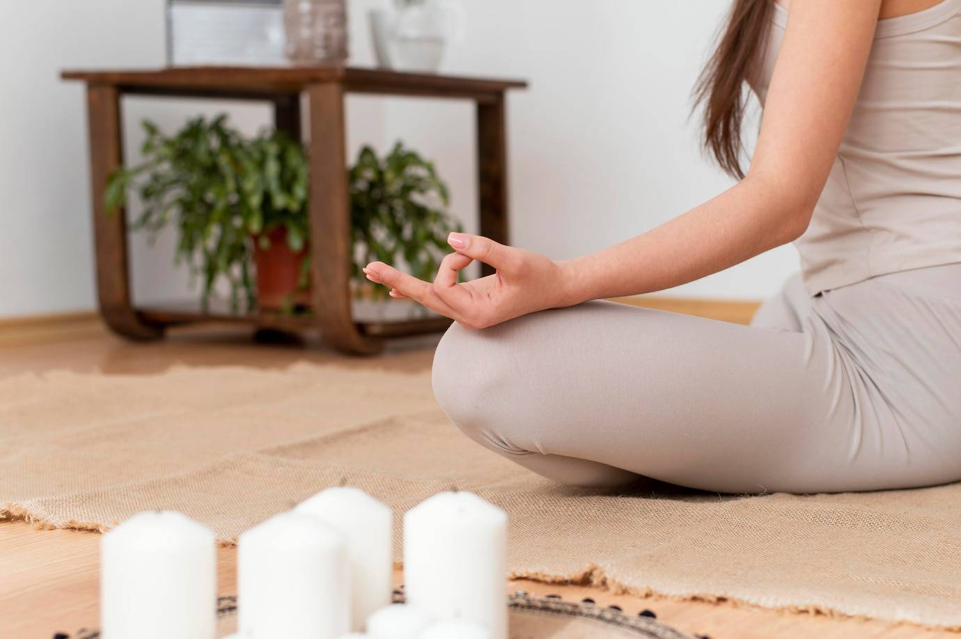 A person meditating in a serene room, seated cross-legged on a natural rug, with lit candles in front and a plant in the background.