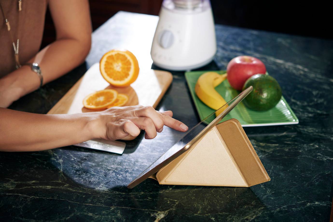 A hand interacts with a tablet on a stand amid fresh fruits and a blender, showcasing a vibrant kitchen scene.