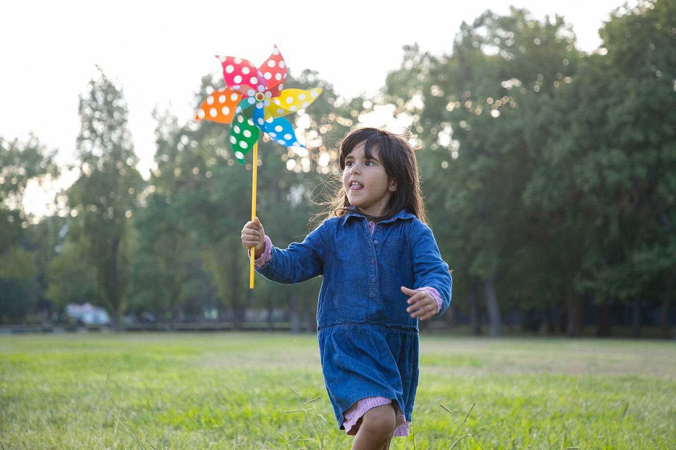 A young child wearing a denim dress joyfully runs in a grassy field, holding a colorful pinwheel. Trees are visible in the background.