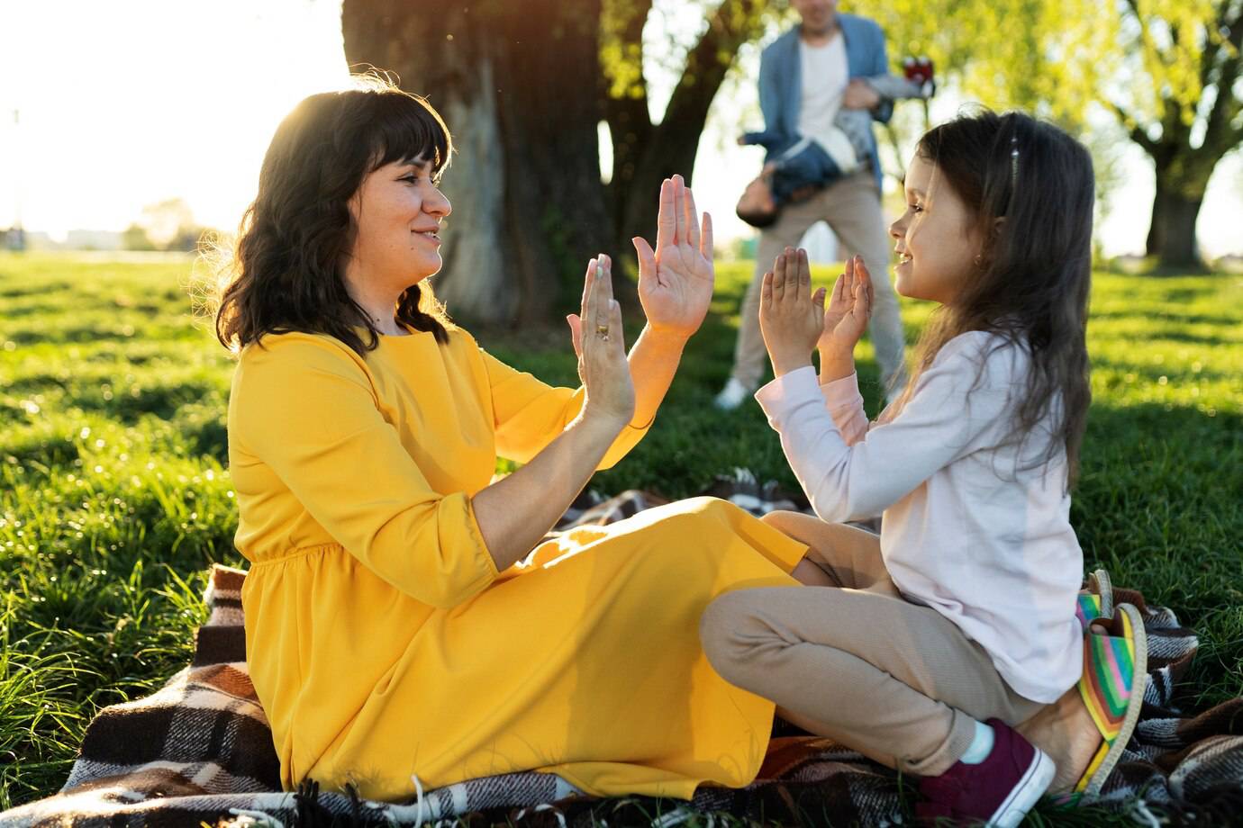 A woman in a yellow dress and a girl play clapping games on a blanket in a sunlit park, surrounded by greenery.
