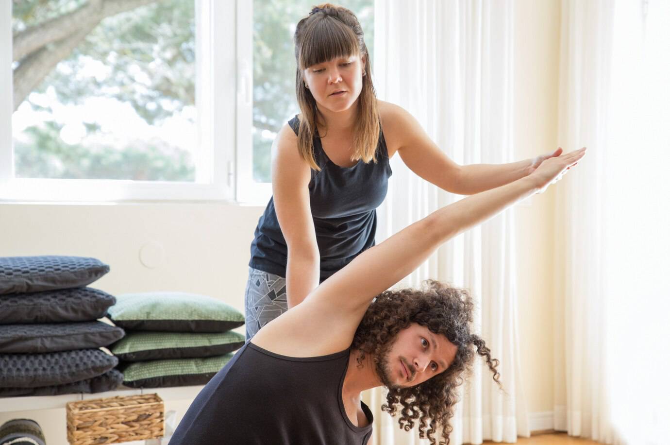 Two women practice stretching in a bright room with large windows. Cushions are neatly stacked nearby, creating a serene atmosphere.