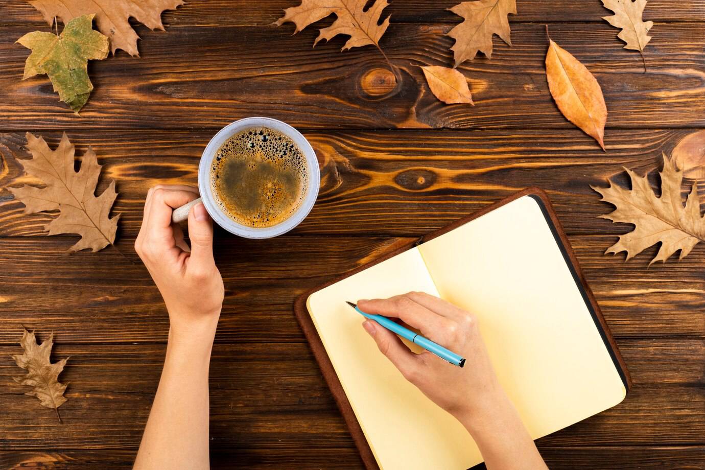 A hand holding a coffee cup beside an open notebook with a blue pen, surrounded by autumn leaves on a wooden table.
