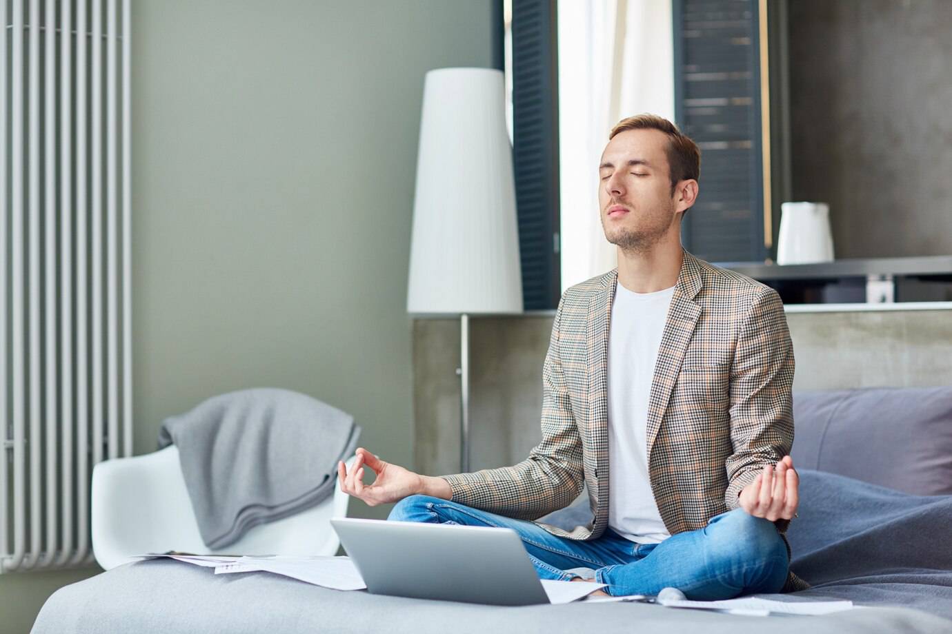 A person sits cross-legged on a bed, meditating with a laptop and papers around in a cozy, modern room.