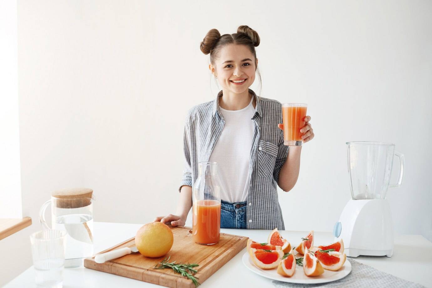 A person in a striped shirt holds a glass of orange juice, surrounded by a blender, a jar of water, and a platter of sliced grapefruit.