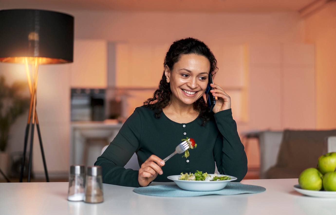 A woman sits at a table, holding a fork with a piece of salad while talking on the phone, surrounded by a cozy home setting.